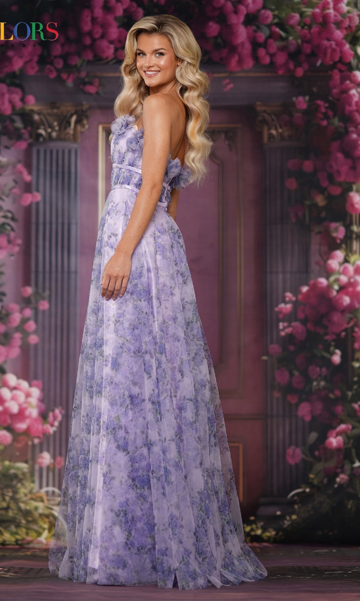 A woman stands sideways in front of a pink and purple floral background, wearing the Colors Dress 3831, a long strapless chiffon prom dress with 3D floral appliques.
